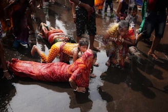 A group of people participate in a ritual involving prostration and prayer in a wet environment. They are wearing colorful traditional clothing with some individuals fully lying on the ground while a woman nearby is in a prayer position. The water on the ground suggests a sacred or ceremonial setting.