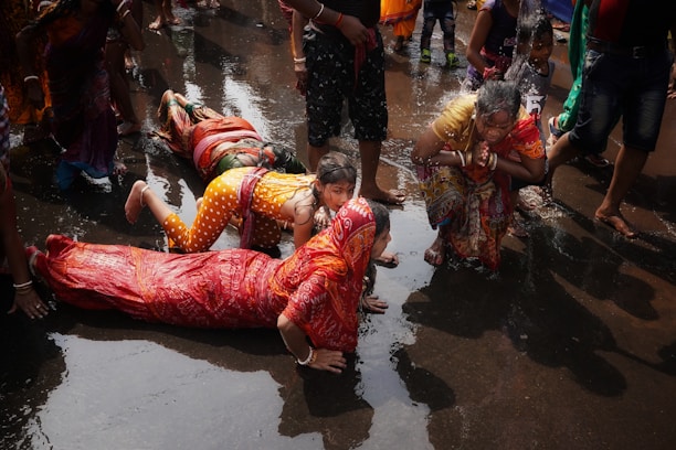 A group of people participate in a ritual involving prostration and prayer in a wet environment. They are wearing colorful traditional clothing with some individuals fully lying on the ground while a woman nearby is in a prayer position. The water on the ground suggests a sacred or ceremonial setting.