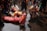 A group of people participate in a ritual involving prostration and prayer in a wet environment. They are wearing colorful traditional clothing with some individuals fully lying on the ground while a woman nearby is in a prayer position. The water on the ground suggests a sacred or ceremonial setting.