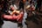 A group of people participate in a ritual involving prostration and prayer in a wet environment. They are wearing colorful traditional clothing with some individuals fully lying on the ground while a woman nearby is in a prayer position. The water on the ground suggests a sacred or ceremonial setting.