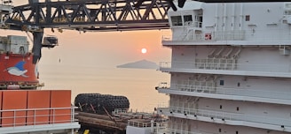 A serene ocean view at sunset with a ship's superstructure on the right. The sun is setting behind an island, casting an orange glow across the sky. Large, round maritime equipment and another ship structure with a dolphin logo are visible in the foreground.