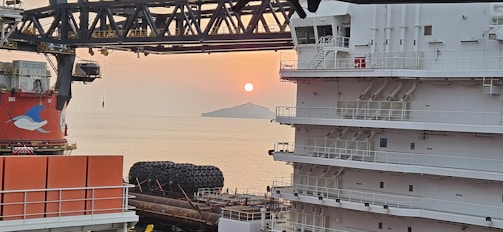 A serene ocean view at sunset with a ship's superstructure on the right. The sun is setting behind an island, casting an orange glow across the sky. Large, round maritime equipment and another ship structure with a dolphin logo are visible in the foreground.