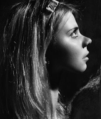 A grayscale profile portrait of a person with long hair, gazing upward. The lighting creates a dramatic contrast, highlighting the contours of the face and the texture of the hair. A decorative hair accessory is visible near the top of the head.