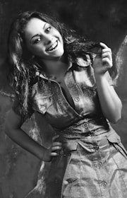 Ethyl smiling brightly, showcasing her 1960s flip hairstyle and mustard-yellow dress in a playful pose.