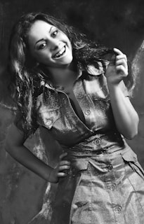 Ethyl smiling brightly, showcasing her 1960s flip hairstyle and mustard-yellow dress in a playful pose.