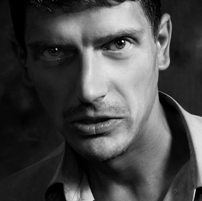 A close-up black and white portrait of a person with a serious expression, featuring sharp facial features, intense eyes, and slight facial hair. The lighting accentuates the contours of the face and creates a dramatic shadow effect.