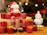 A decorative Santa figurine sits atop a wooden block surrounded by multiple small, glittery red gift boxes with yellow ribbons. Next to Santa stands a snowman figure wearing a red hat and scarf. The background features blurred Christmas lights and a tree.