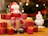 A decorative Santa figurine sits atop a wooden block surrounded by multiple small, glittery red gift boxes with yellow ribbons. Next to Santa stands a snowman figure wearing a red hat and scarf. The background features blurred Christmas lights and a tree.