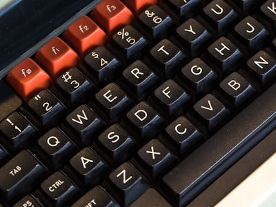 A close-up view of a mechanical keyboard featuring black keycaps with white lettering. The top row has red keycaps with white lettering, representing function keys. The layout is QWERTY with special symbols and numbers combined on some keys.