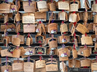 A collection of wooden plaques with various handwritten messages, some decorated with illustrations. The plaques are hung with colorful strings on a wooden structure, creating an arrangement of wishes or prayers.