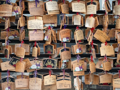 A collection of wooden plaques with various handwritten messages, some decorated with illustrations. The plaques are hung with colorful strings on a wooden structure, creating an arrangement of wishes or prayers.