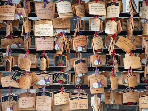 A collection of wooden plaques with various handwritten messages, some decorated with illustrations. The plaques are hung with colorful strings on a wooden structure, creating an arrangement of wishes or prayers.