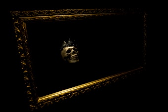A human skull crowned with a dark, possibly black, crown is displayed within an ornate golden frame against a black background. The skull is the central focus, illuminated subtly to emphasize its features amidst the surrounding darkness.
