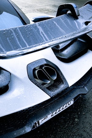 A close-up view of the rear of a sports car featuring a large spoiler and dual exhaust pipes. The surface is covered in water droplets, giving it a sleek and freshly washed appearance. The license plate is visible at the bottom, with a mix of numbers and Arabic text.