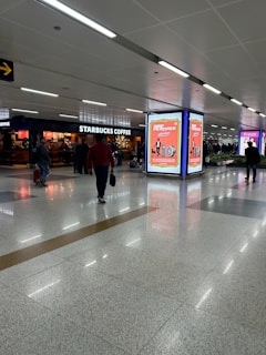 An airport terminal with people walking by, featuring a Starbucks Coffee shop on one side. There are illuminated advertisement panels with red and white designs, and the floor is a polished tile surface. The ceiling has recessed lighting.