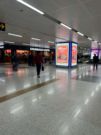 An airport terminal with people walking by, featuring a Starbucks Coffee shop on one side. There are illuminated advertisement panels with red and white designs, and the floor is a polished tile surface. The ceiling has recessed lighting.