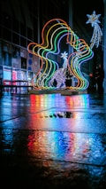 A vibrant and colorful light installation is displayed on a wet pavement, reflecting vivid hues onto the ground. The lights are arranged in an artistic shape and accompanied by other illuminated decorations, such as stars. People can be seen in the background, adding a sense of scale and context to the urban setting.