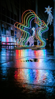 A vibrant and colorful light installation is displayed on a wet pavement, reflecting vivid hues onto the ground. The lights are arranged in an artistic shape and accompanied by other illuminated decorations, such as stars. People can be seen in the background, adding a sense of scale and context to the urban setting.