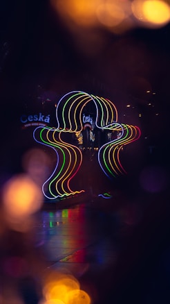A vivid neon sign displays colorful, wavy vertical lines in a dark setting, reflecting on a wet or polished surface. The word 'Cesk&aacute;' is visible, suggesting a possible association with a Czech-themed location or establishment. The background is blurred, with soft circles of light providing a bokeh effect.