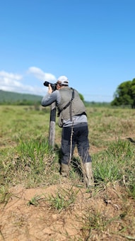 A person wearing outdoor clothing and boots is standing in a grassy field, holding a camera with a long lens, possibly aimed at wildlife or a landscape. The sky is clear and blue, with some clouds in the background, and there are trees visible in the distance.