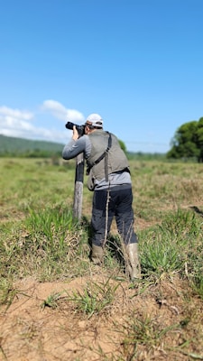 A person wearing outdoor clothing and boots is standing in a grassy field, holding a camera with a long lens, possibly aimed at wildlife or a landscape. The sky is clear and blue, with some clouds in the background, and there are trees visible in the distance.