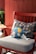 A blue plush toy featuring a smiling face and colorful patches of yellow, green, and white rests on a light gray cushion of a red wooden chair. Behind the toy is a black pillow with geometric patterns in various colors. The background has a reddish paneled wall. Partially visible to the left are green plant leaves.