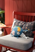 A blue plush toy featuring a smiling face and colorful patches of yellow, green, and white rests on a light gray cushion of a red wooden chair. Behind the toy is a black pillow with geometric patterns in various colors. The background has a reddish paneled wall. Partially visible to the left are green plant leaves.