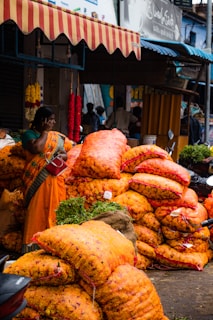 A bustling market scene with a woman wearing a colorful sari standing near large sacks of bright orange marigolds. The sacks are stacked high, suggesting a wholesale trading environment. Hanging flower garlands add to the lively atmosphere, while motorcycles and people in the background indicate a busy street setting.