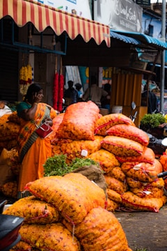 A bustling market scene with a woman wearing a colorful sari standing near large sacks of bright orange marigolds. The sacks are stacked high, suggesting a wholesale trading environment. Hanging flower garlands add to the lively atmosphere, while motorcycles and people in the background indicate a busy street setting.