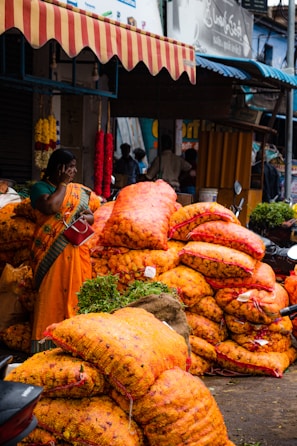 A bustling market scene with a woman wearing a colorful sari standing near large sacks of bright orange marigolds. The sacks are stacked high, suggesting a wholesale trading environment. Hanging flower garlands add to the lively atmosphere, while motorcycles and people in the background indicate a busy street setting.
