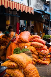 A bustling market scene with a woman wearing a colorful sari standing near large sacks of bright orange marigolds. The sacks are stacked high, suggesting a wholesale trading environment. Hanging flower garlands add to the lively atmosphere, while motorcycles and people in the background indicate a busy street setting.