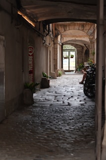 A narrow, cobbled pathway leads through an archway surrounded by old stone buildings. A bicycle rental sign is visible on the left, accompanied by a row of bicycles. Green plants are seen decorating the pathway, and natural light pours through an open gate at the far end. A person is seated near the bikes, adding a casual touch to the scene.