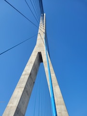 A modern cable-stayed bridge tower extending upwards with a strong geometric design. The structure features vertical cables and a tall concrete pillar against a clear blue sky.