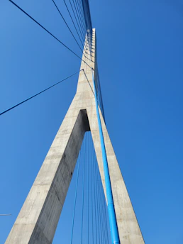 A modern cable-stayed bridge tower extending upwards with a strong geometric design. The structure features vertical cables and a tall concrete pillar against a clear blue sky.