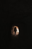A person is walking through a dark tunnel toward the light at the end. The silhouette is visible against the brighter outdoors, suggesting a transition from darkness to light.