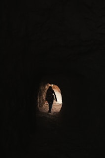 A person is walking through a dark tunnel toward the light at the end. The silhouette is visible against the brighter outdoors, suggesting a transition from darkness to light.