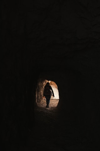 A person is walking through a dark tunnel toward the light at the end. The silhouette is visible against the brighter outdoors, suggesting a transition from darkness to light.