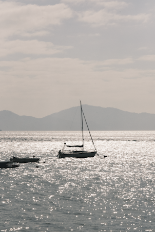 A sailboat floats on a shimmering, sunlit sea with soft waves. In the background, misty mountains create a tranquil horizon under a partially cloudy sky.