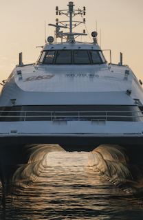 A close-up of the front view of a large, modern catamaran-style ship with a white body and windows facing forward. The reflection of the sun creates a golden glow on the water beneath the ship. The structure above the deck includes antennas and lights.