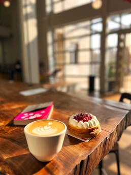 A wooden table with a white ceramic cup containing a latte, topped with latte art, sits in a warmly lit cafe. Next to the cup is a pastry adorned with cream and pomegranate seeds. In the background, a pink book is partially visible. The cafe has large, sunlit windows creating a cozy atmosphere.