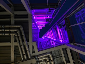 A modern architectural interior featuring a series of metallic pipes running parallel along a wall, illuminated by vivid purple lighting. The perspective creates a sense of depth with geometric patterns and a play of shadows.