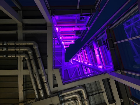 A modern architectural interior featuring a series of metallic pipes running parallel along a wall, illuminated by vivid purple lighting. The perspective creates a sense of depth with geometric patterns and a play of shadows.