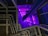 A modern architectural interior featuring a series of metallic pipes running parallel along a wall, illuminated by vivid purple lighting. The perspective creates a sense of depth with geometric patterns and a play of shadows.