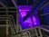 A modern architectural interior featuring a series of metallic pipes running parallel along a wall, illuminated by vivid purple lighting. The perspective creates a sense of depth with geometric patterns and a play of shadows.