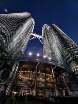 Two tall, modern skyscrapers illuminated against the night sky connected by a skybridge near the top. The buildings have glass facades with a metallic sheen, reflecting the lights. At the base, large entrance doors and surrounding landscape are visible, as well as silhouettes of people.