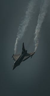 A fighter jet is captured mid-flight against a dark sky, with trails of vapor streaming from its wings as it maneuvers through the air.