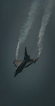A fighter jet is captured mid-flight against a dark sky, with trails of vapor streaming from its wings as it maneuvers through the air.