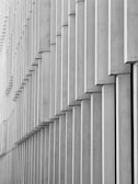 Geometric patterns created by a series of vertical concrete slabs arranged in a staggered formation. The slabs cast shadows, resulting in a layered and textured appearance.
