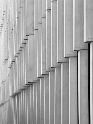 Geometric patterns created by a series of vertical concrete slabs arranged in a staggered formation. The slabs cast shadows, resulting in a layered and textured appearance.