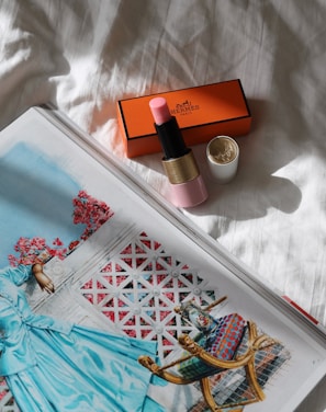 A pink lipstick with a gold and black tube lies next to its opened white cap on a white textured fabric. Nearby, an orange Hermès box is placed, partially shaded. An open magazine displaying an illustration of a person in a blue garment leaning on a decorative balcony is also visible, with a vibrant color palette including red flowers and a geometric background.